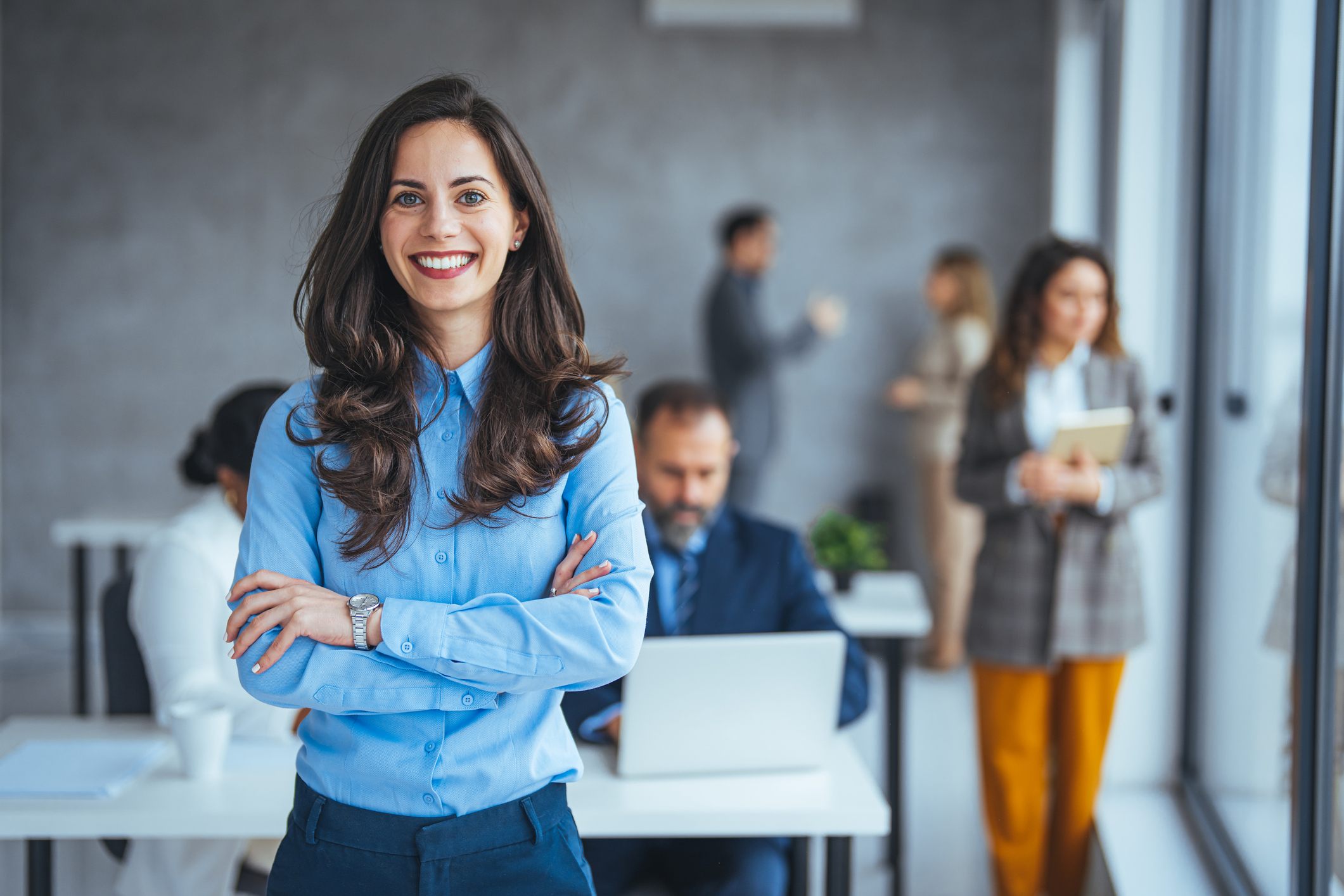 Professional woman in a blue shirt smiling with arms crossed in a modern office, colleagues working and talking in the background.